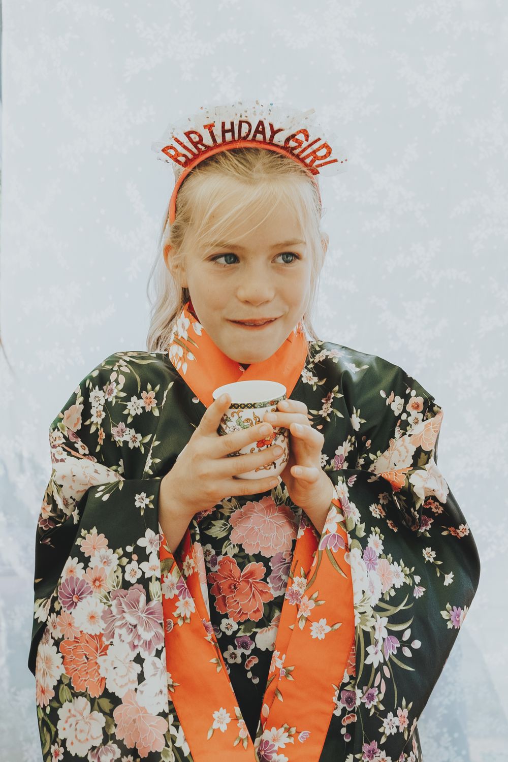 A young birthday girl wearing a floral kimono and Birthday Girl tiara holds a party cup.