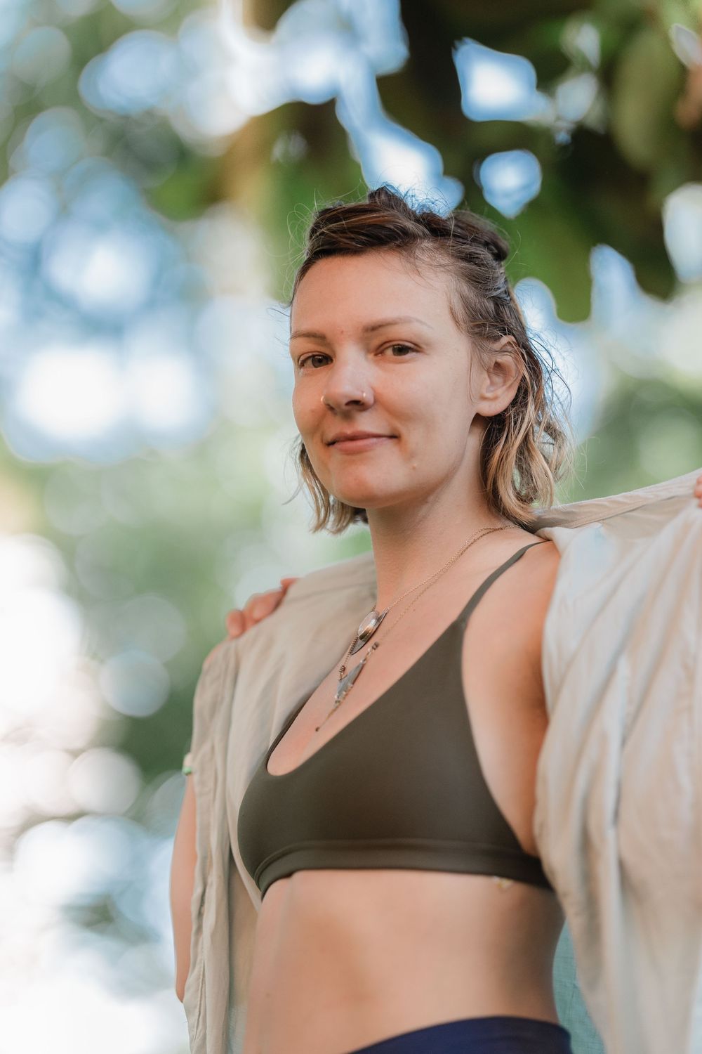 Young woman in olive green sports bra smiling softly under lush green trees outdoors.