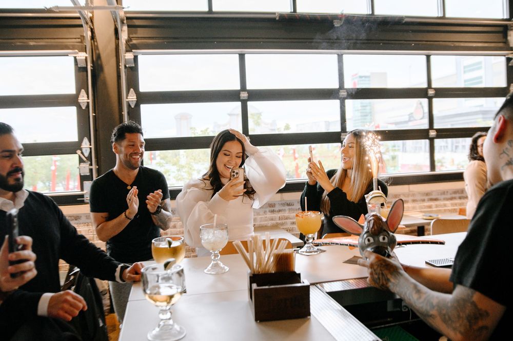 Group of people enjoying drinks and conversation at a long table in a bright restaurant setting.