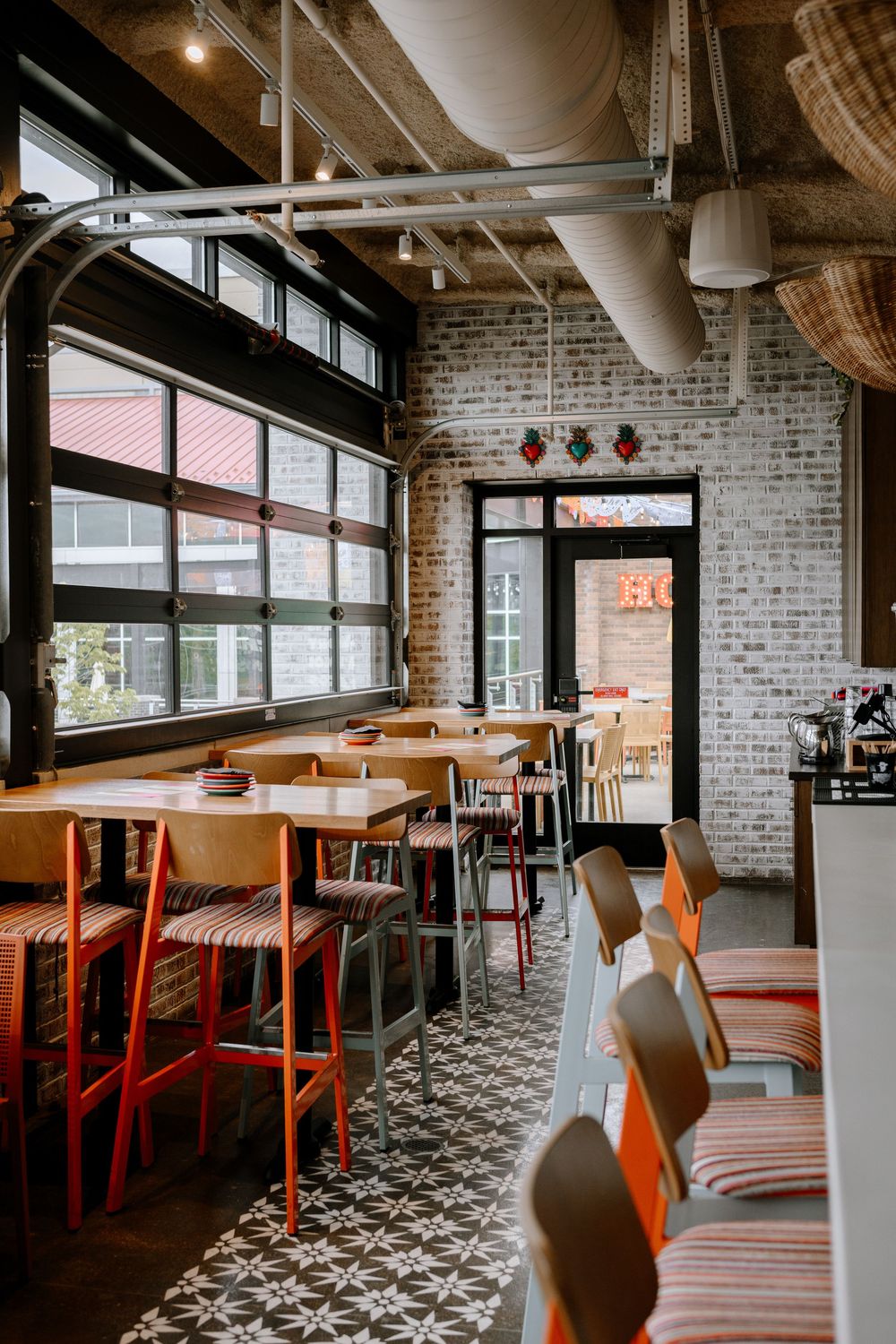 Modern restaurant interior with patterned floor tiles, orange chairs, and exposed brick walls illuminated by pendant lighting.