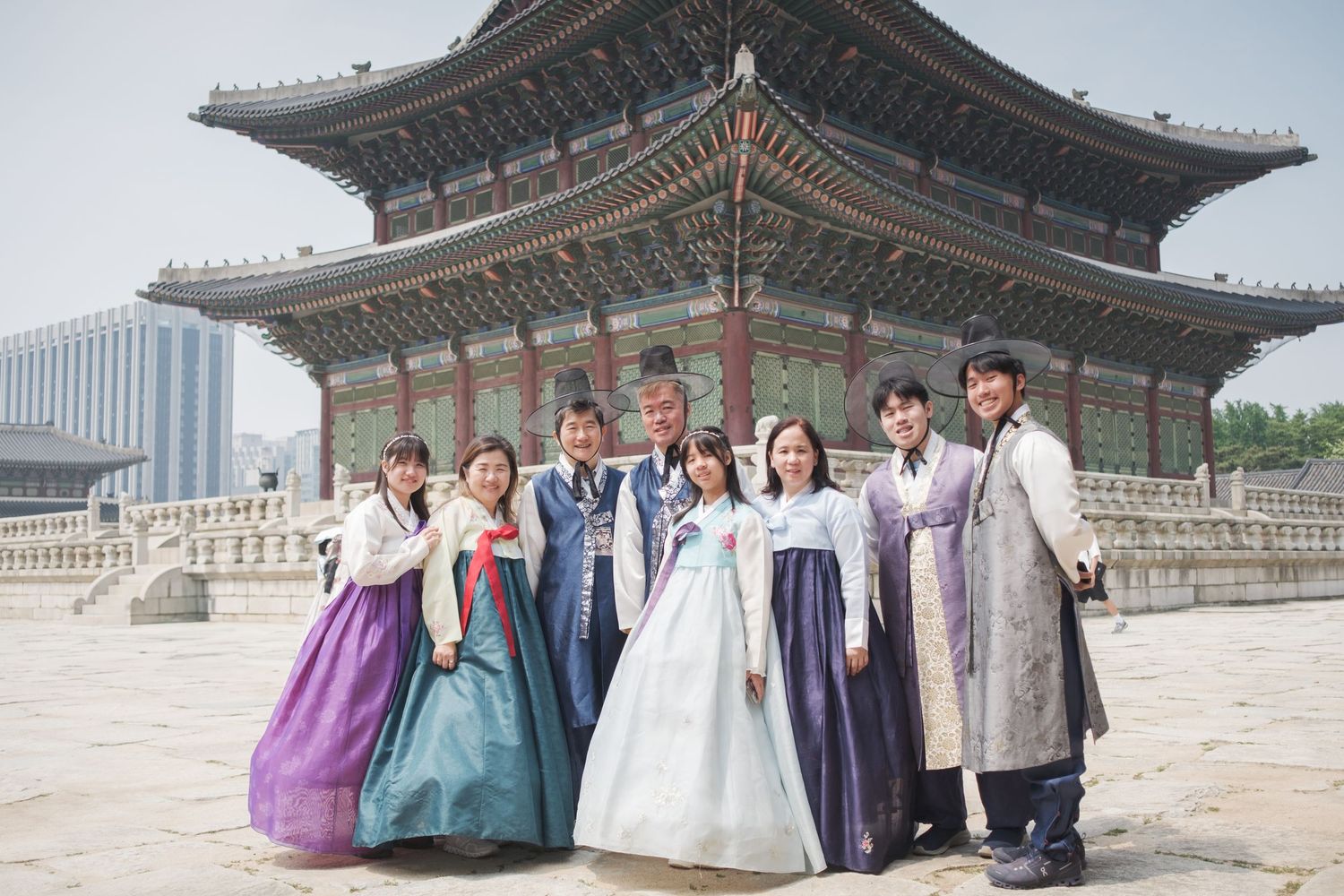 Group wearing traditional Korean hanbok clothing poses in front of historic Gyeongbokgung Palace architecture in Seoul.