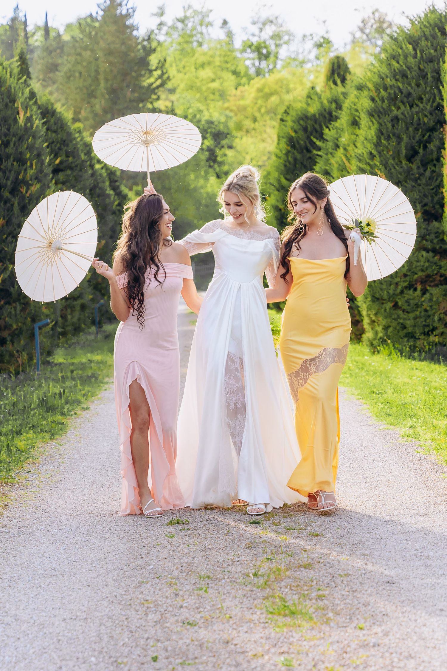 Bridal party in pastel dresses walks along a path holding white parasols.