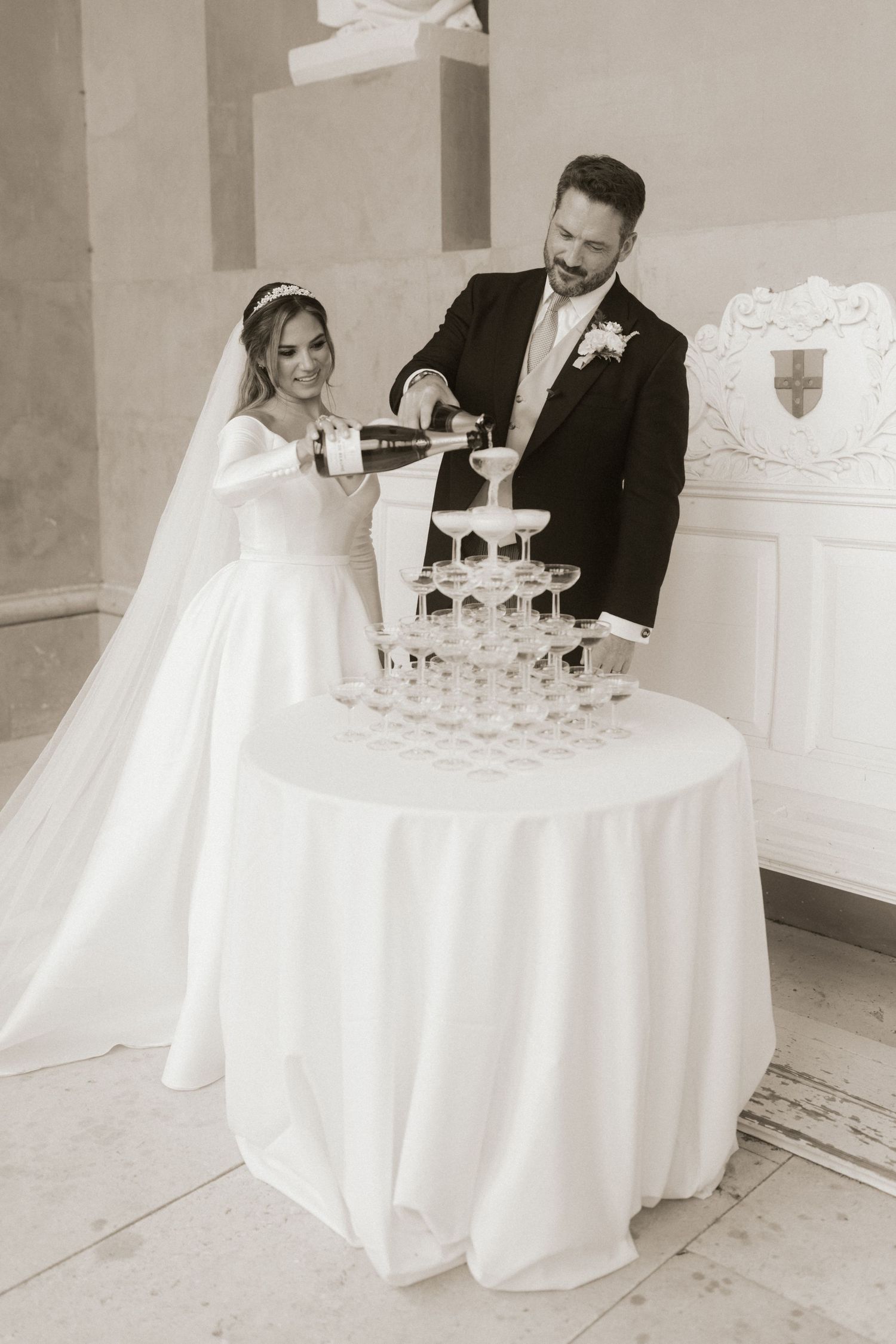 Wedding couple pouring champagne into a tower of glasses at their elegant white reception.