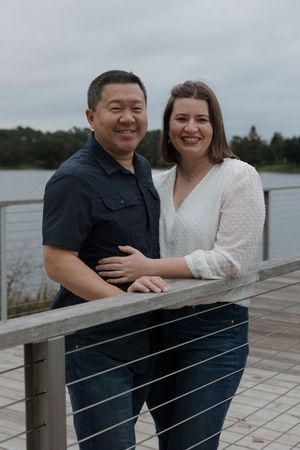 A couple leans against a wooden railing overlooking a lake on an overcast day.