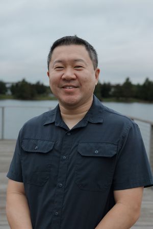 A person in a black button-up shirt smiles warmly at the camera outdoors.
