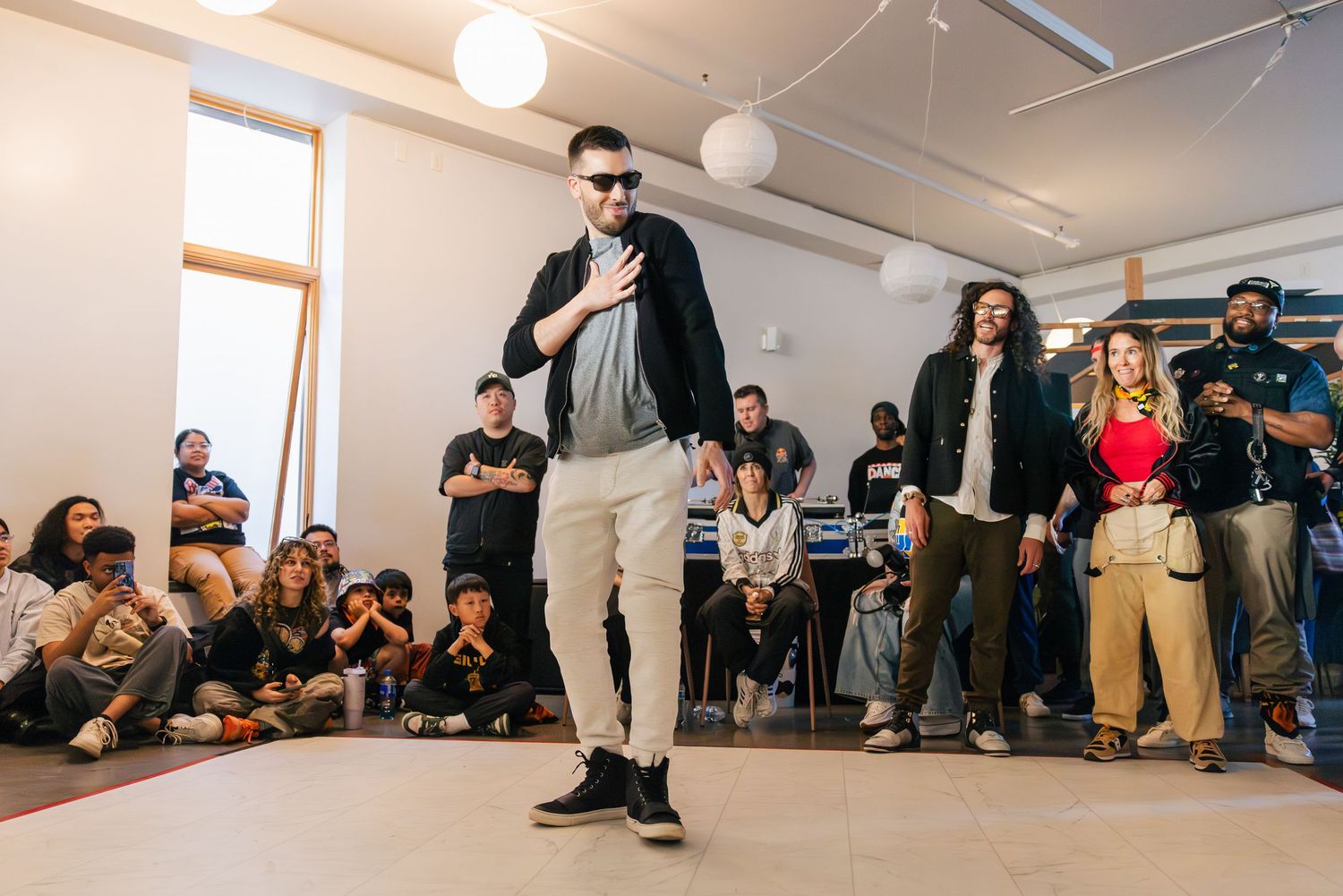People sit and stand along the walls of a bright white gallery or studio space during what appears to be an event.