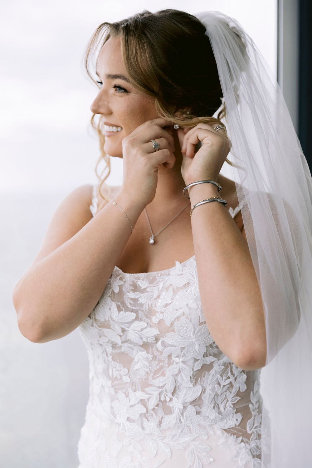 Bride putting on earrings while wearing lace detailed wedding dress.