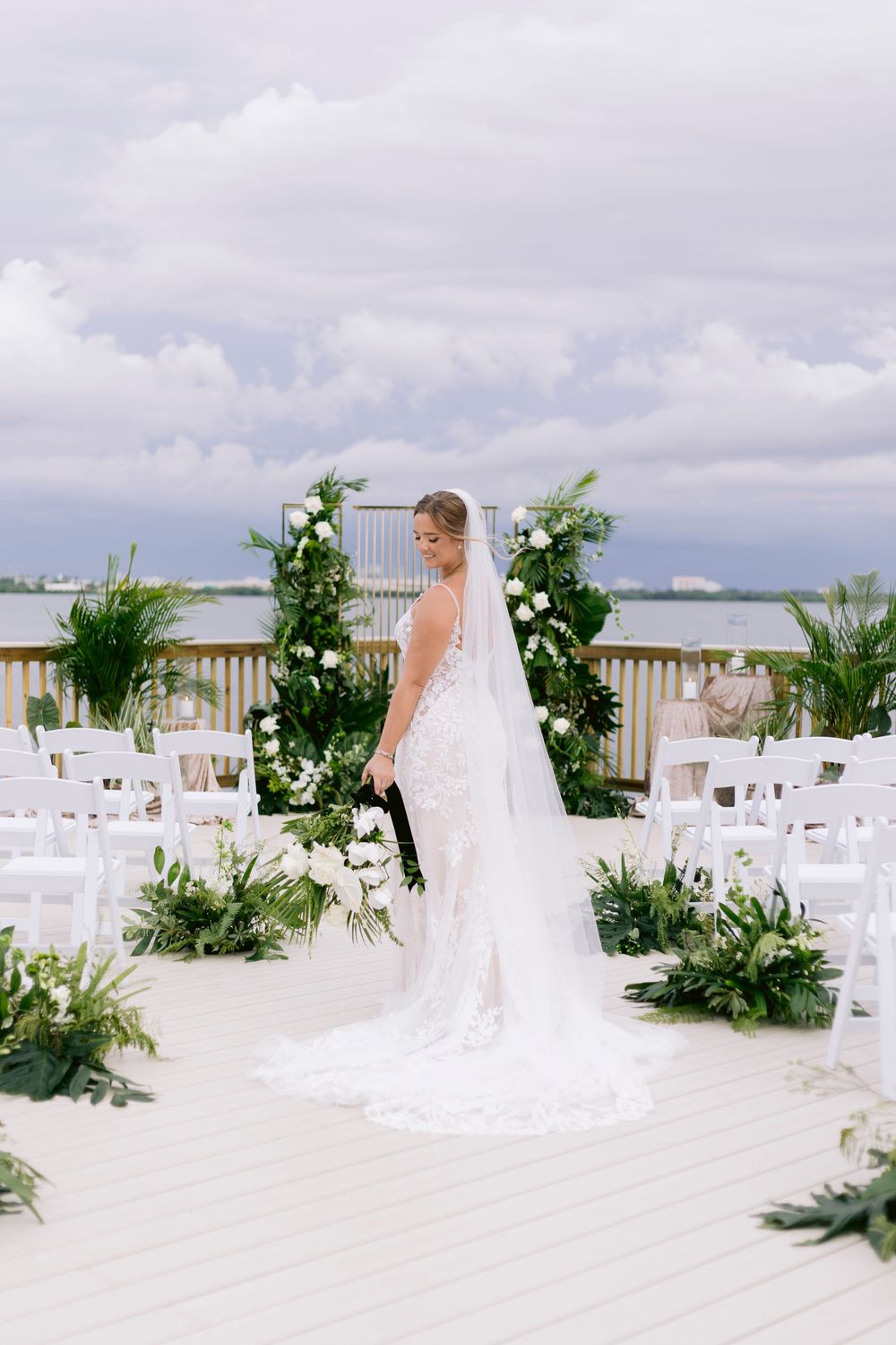 White chairs and tropical greenery decorate an outdoor waterfront wedding ceremony setup.