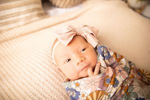 A baby lies on beige bedding while wearing a floral dress and matching headband.