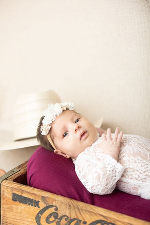 Baby in white lace dress lying on vintage wooden Coca-Cola crate in artistic portrait series.