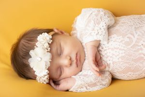 Sleeping baby in white lace dress and floral headband lying on yellow backdrop during professional photo session.