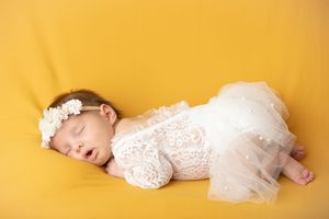 A baby sleeping peacefully in a white lace dress against a bright yellow background.