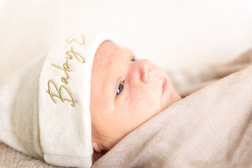 A newborn baby wearing a white knit cap lies peacefully on soft white bedding.