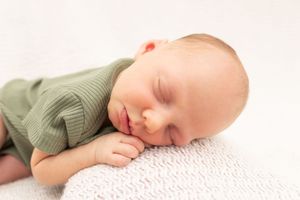 A sleeping baby in a sage green outfit rests peacefully on white bedding.
