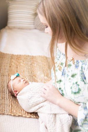 A newborn baby in white knit clothing lies on a textured beige rug while being gently cared for in soft natural light.
