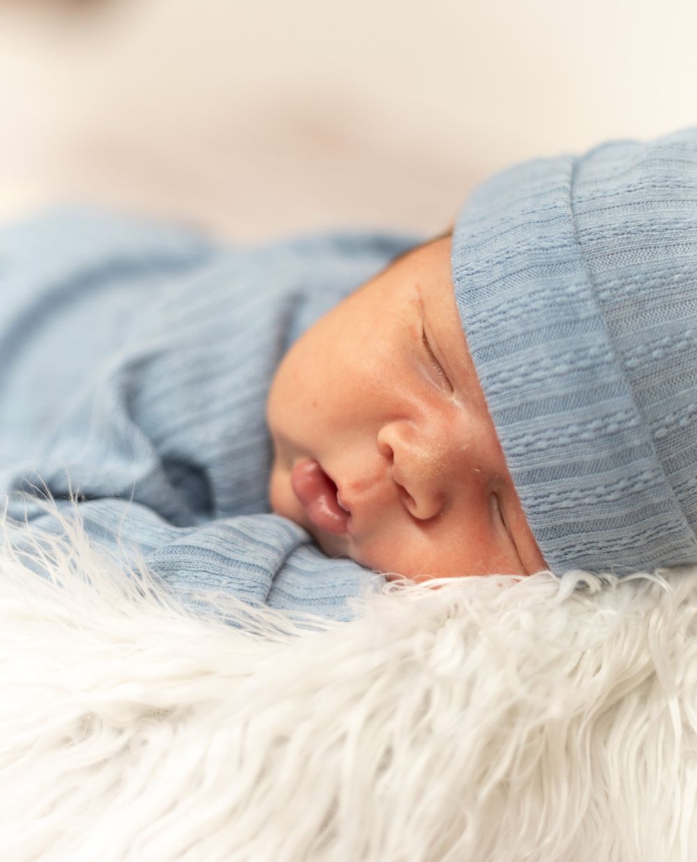 A sleeping baby wrapped in a cozy gray blanket rests peacefully on white fluffy fur bedding.