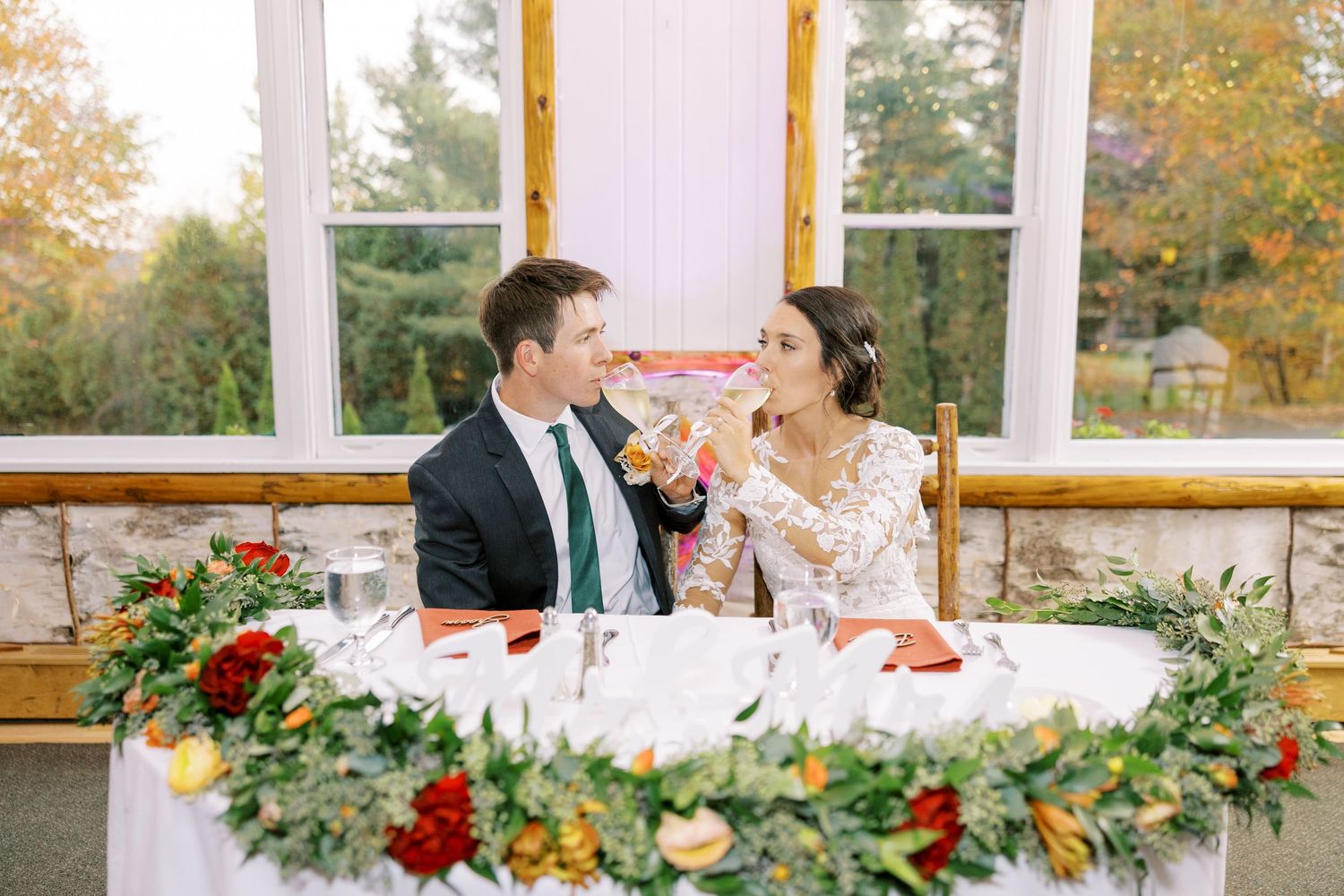 Newlyweds share an intimate moment at their wedding reception table decorated with lush greenery and red roses.