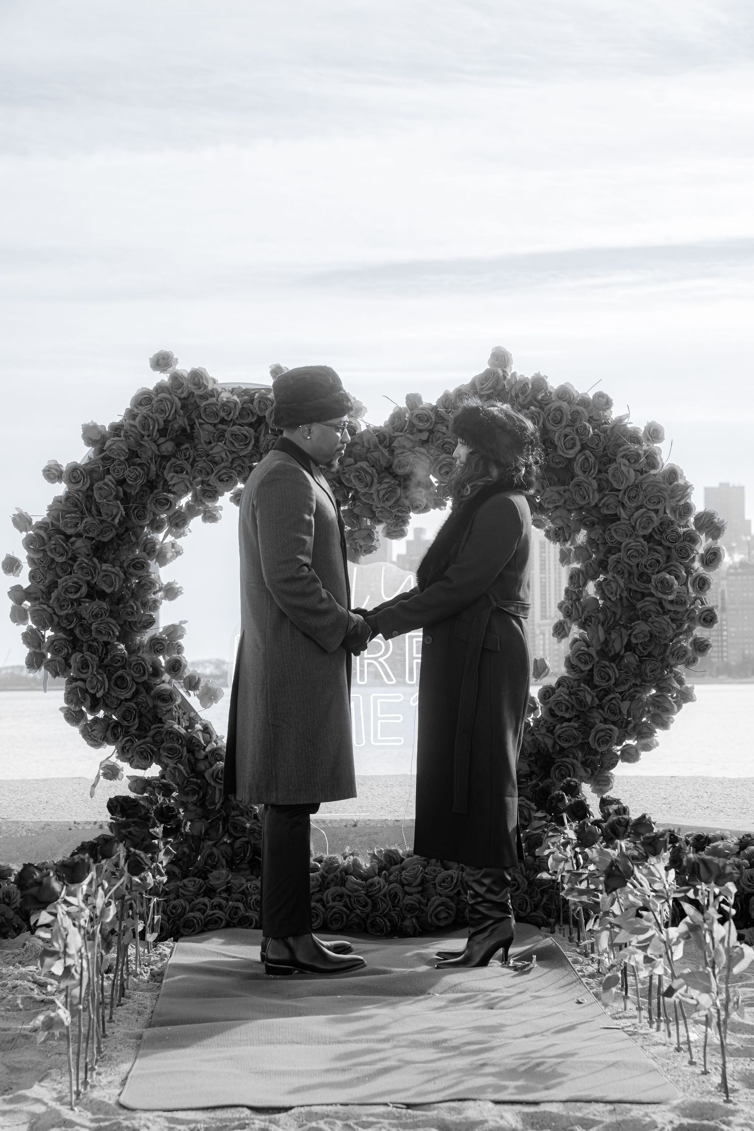 A vintage black and white romantic scene shows a couple under a floral heart-shaped arch during a wedding ceremony.
