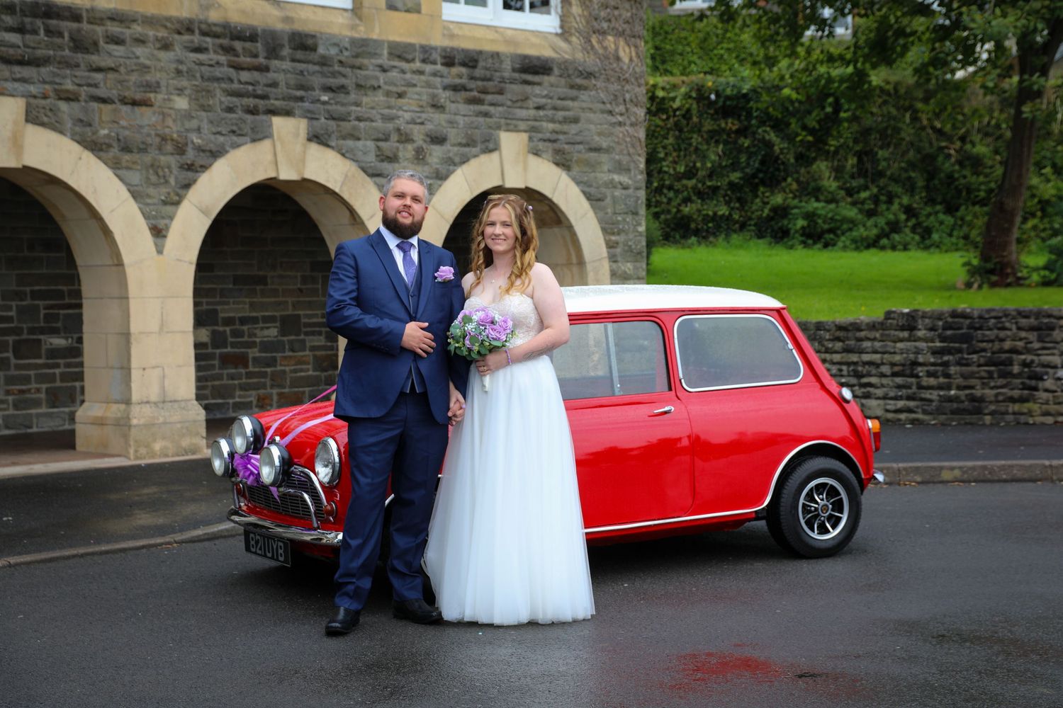 Wedding couple poses beside classic red Mini Cooper outside stone building.