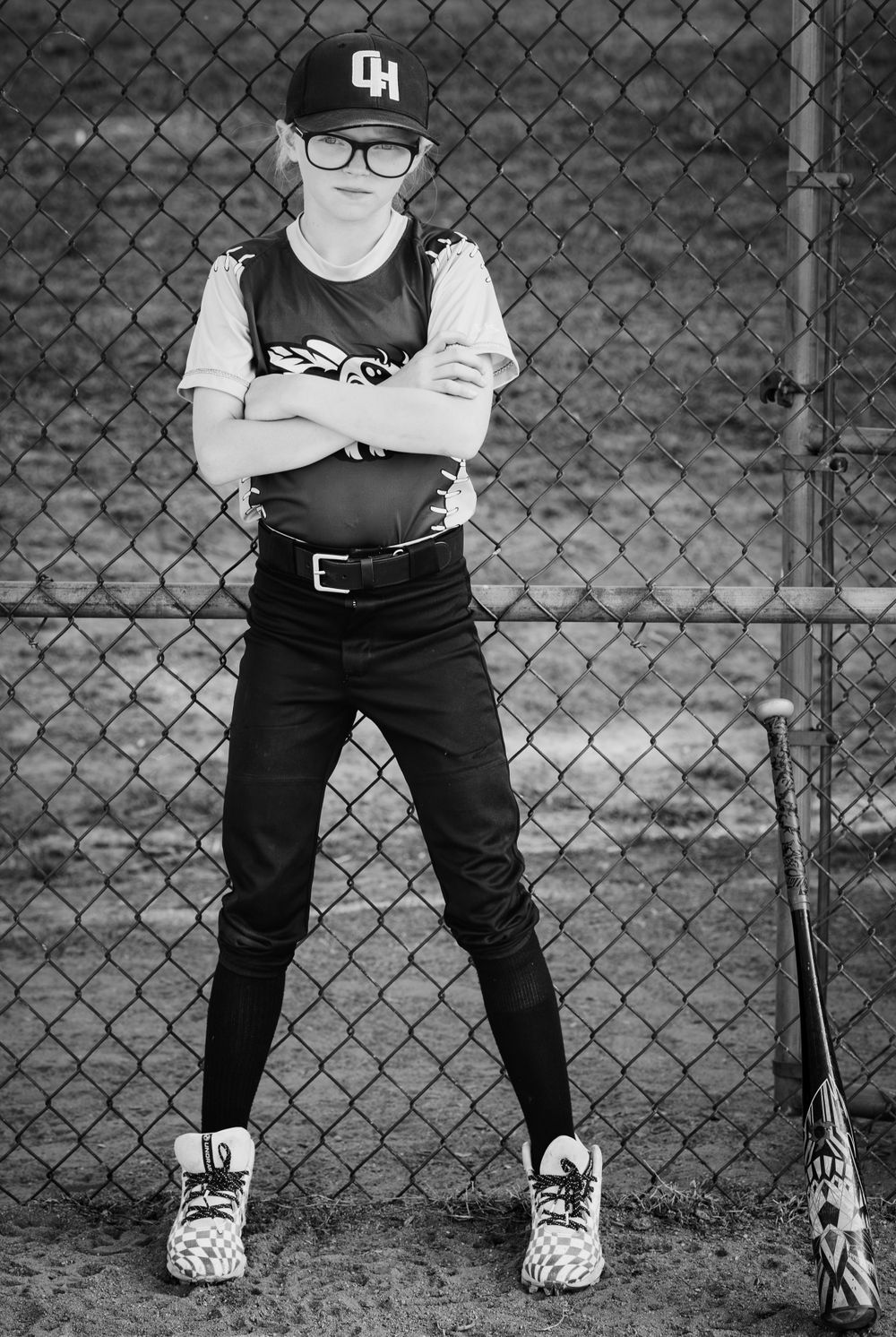 Black and white portrait of a young baseball player in glasses and cap standing against a chain link fence.