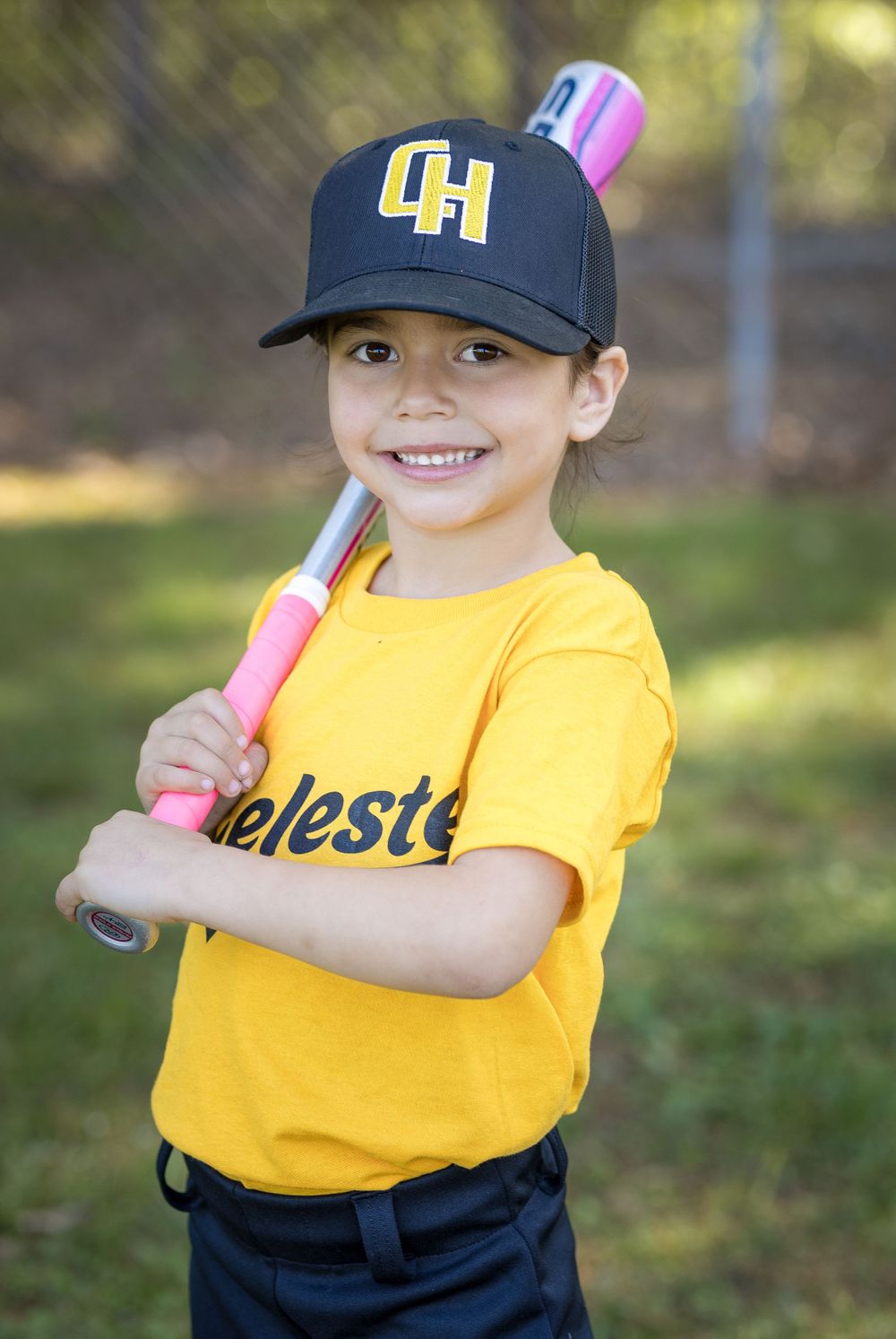 Young baseball player in yellow jersey and navy cap holding pink bat in sunny outdoor setting.