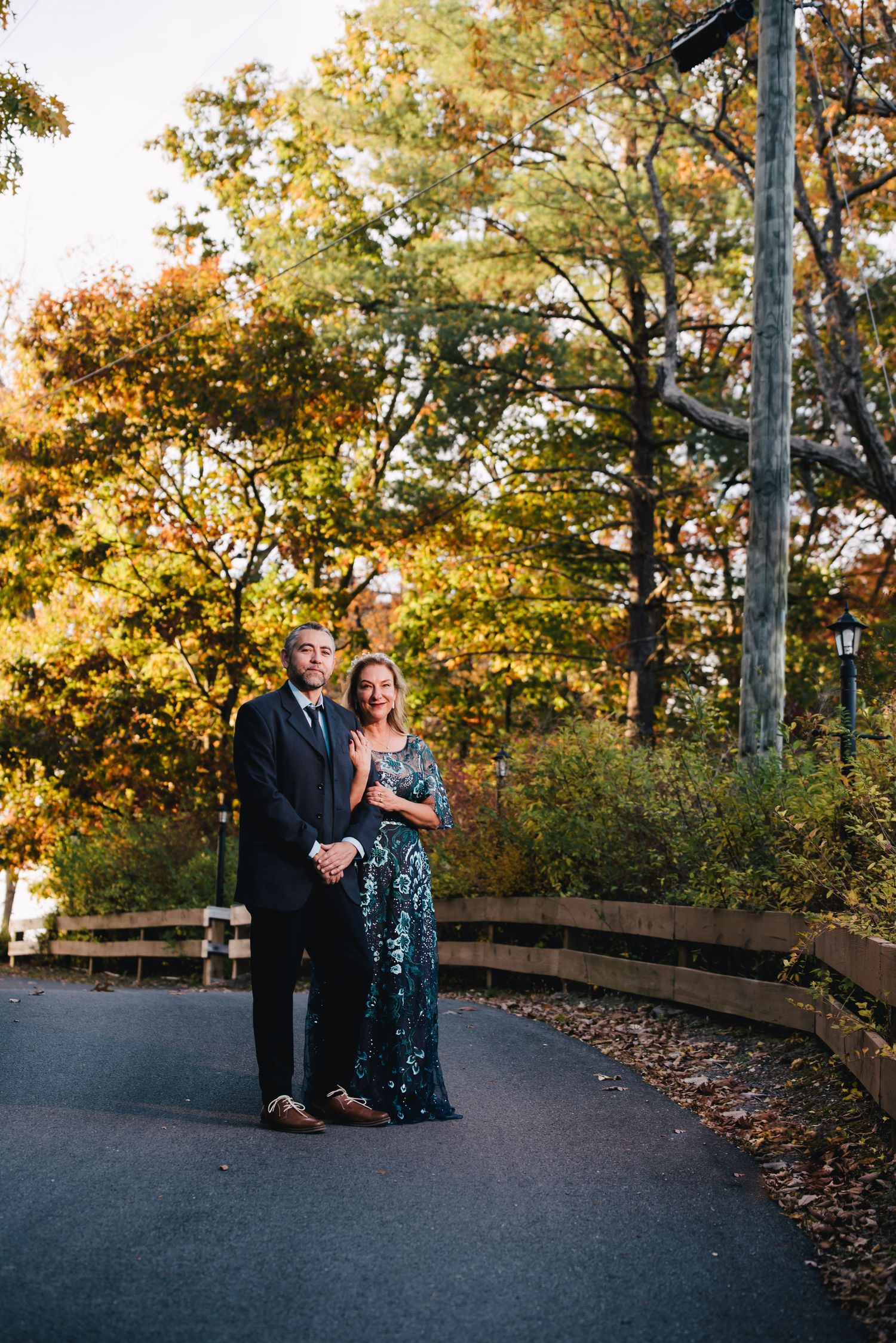 A formal couple poses on a curved path during golden hour with autumn foliage in the background.