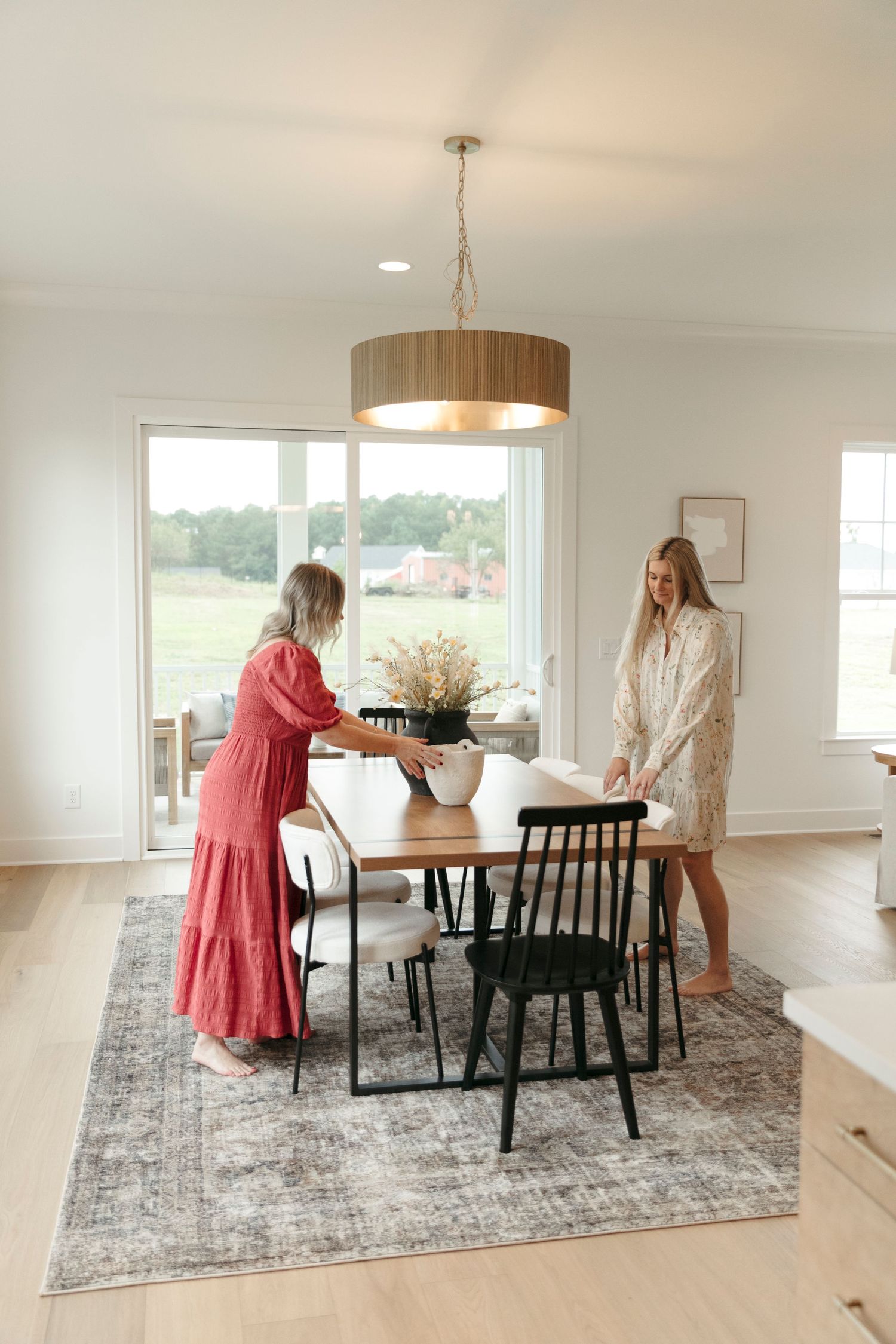 Modern dining room with wooden drum pendant light, round table, and two people setting up flowers in a bright space.