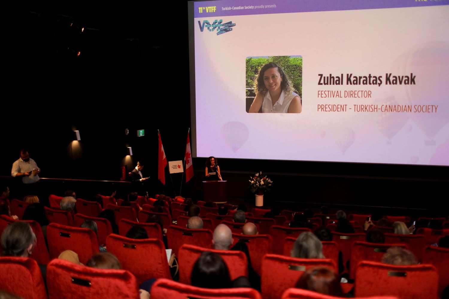A theater audience sits in red seats facing a stage with a projection screen displaying a presentation slide.