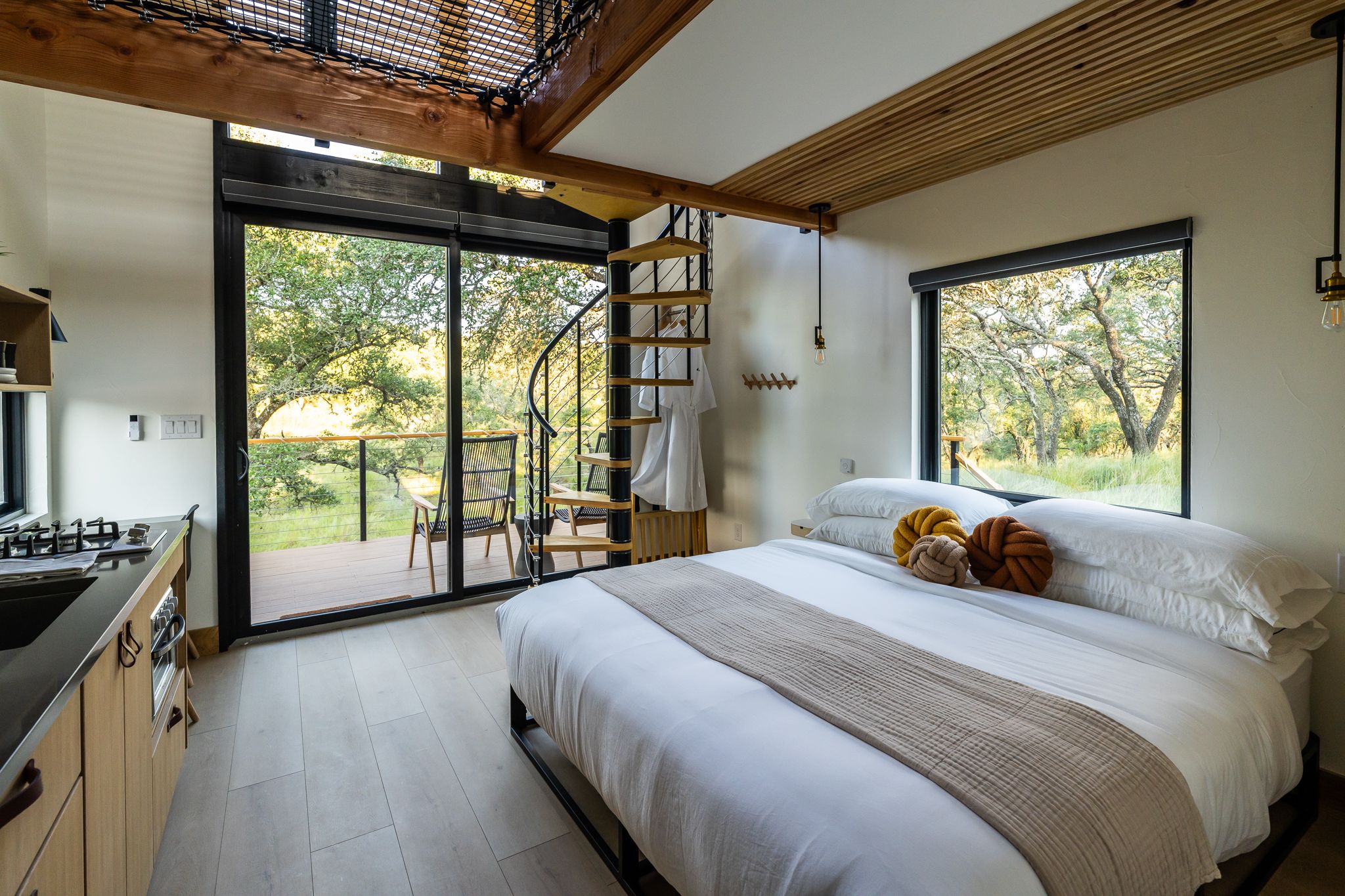 Modern cabin bedroom with large windows, spiral staircase, and natural light flooding the space.