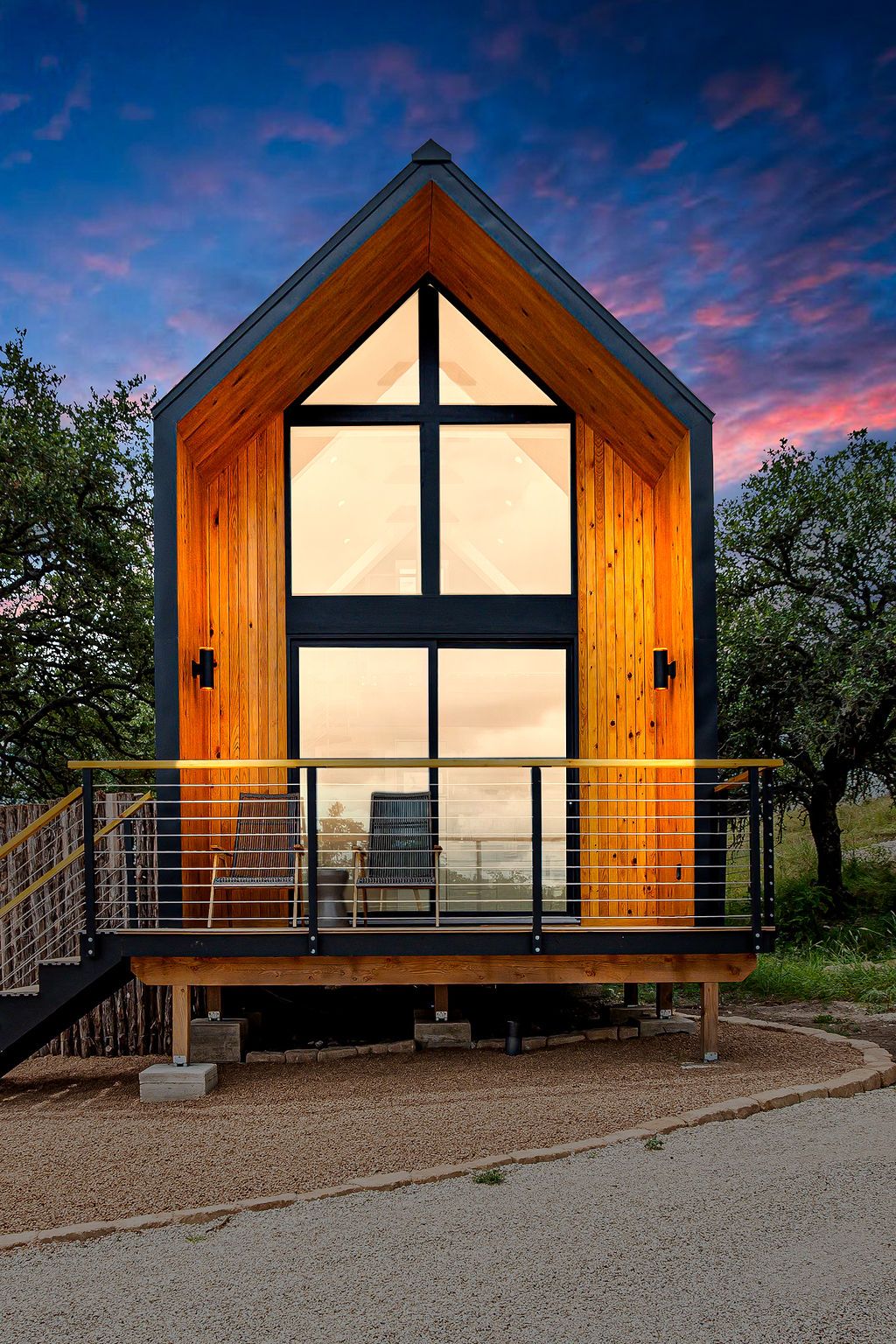 Modern A-frame cabin with large glass windows glows warmly against a dramatic sunset sky.
