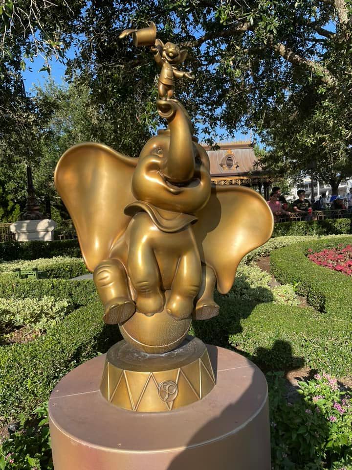A golden statue of Dumbo the elephant balancing on a circus platform in a Disney park garden setting.