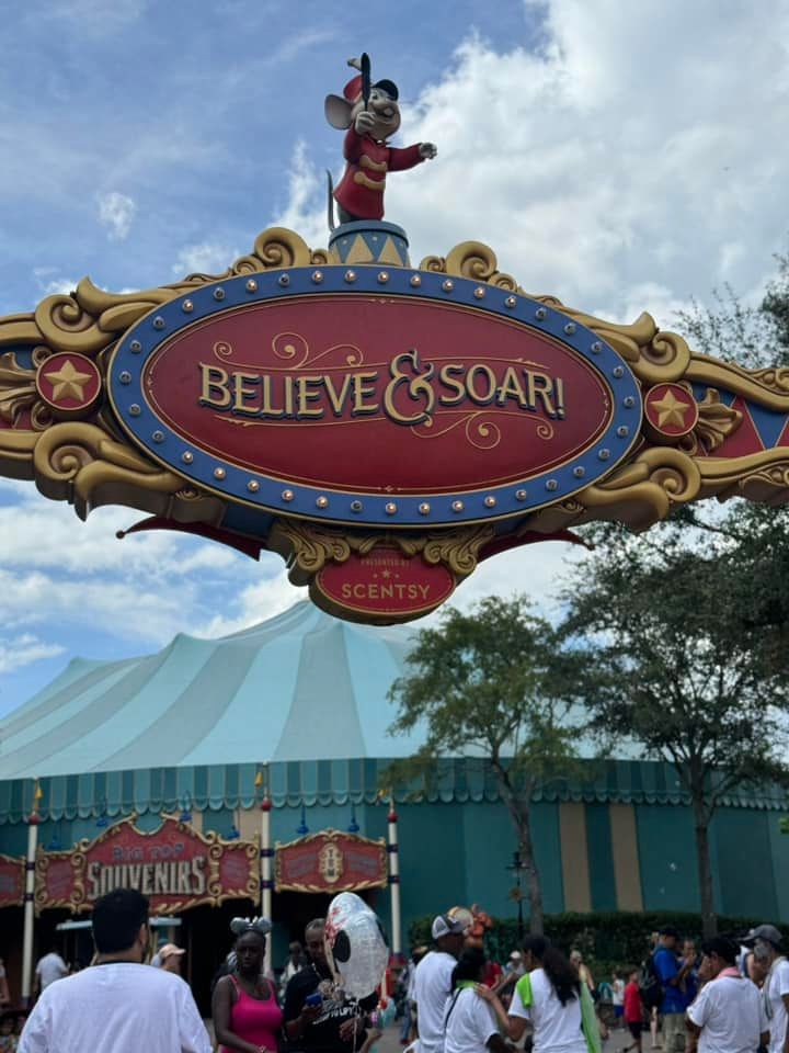 A colorful theme park sign reading 'Believe & Soar!' with decorative ornaments against a blue sky at an amusement park entrance.