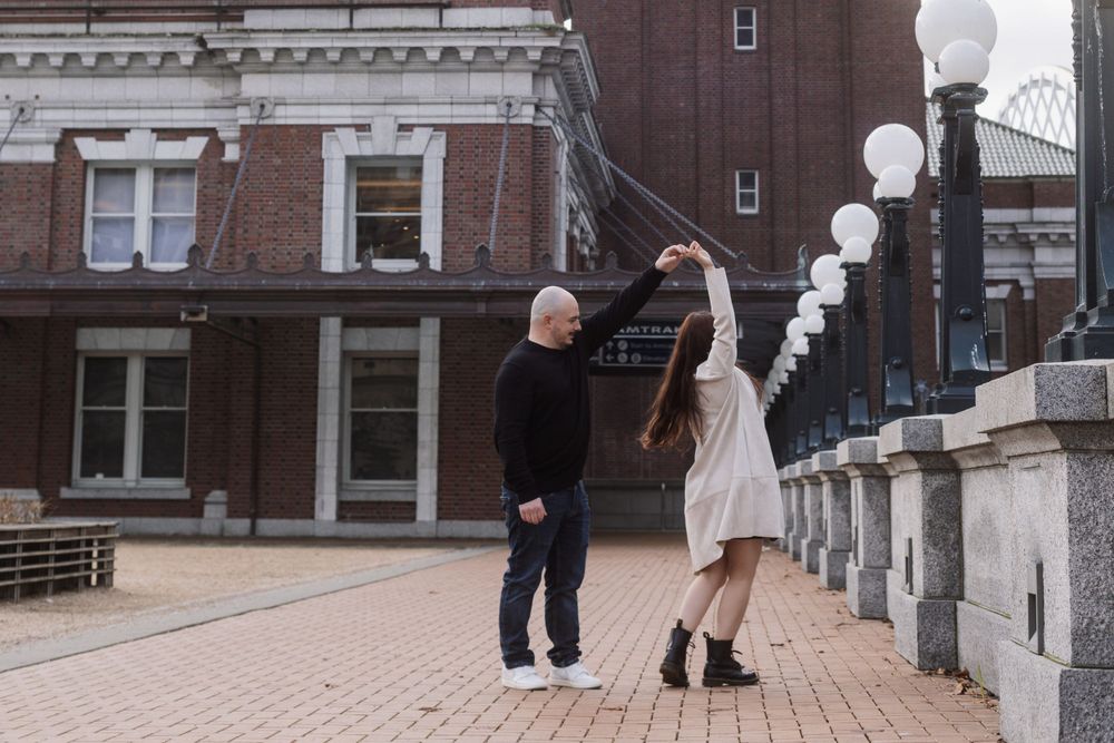 Happy couple dressed casually dance together on a brick terrace with white lamp posts and historic building backdrop.
