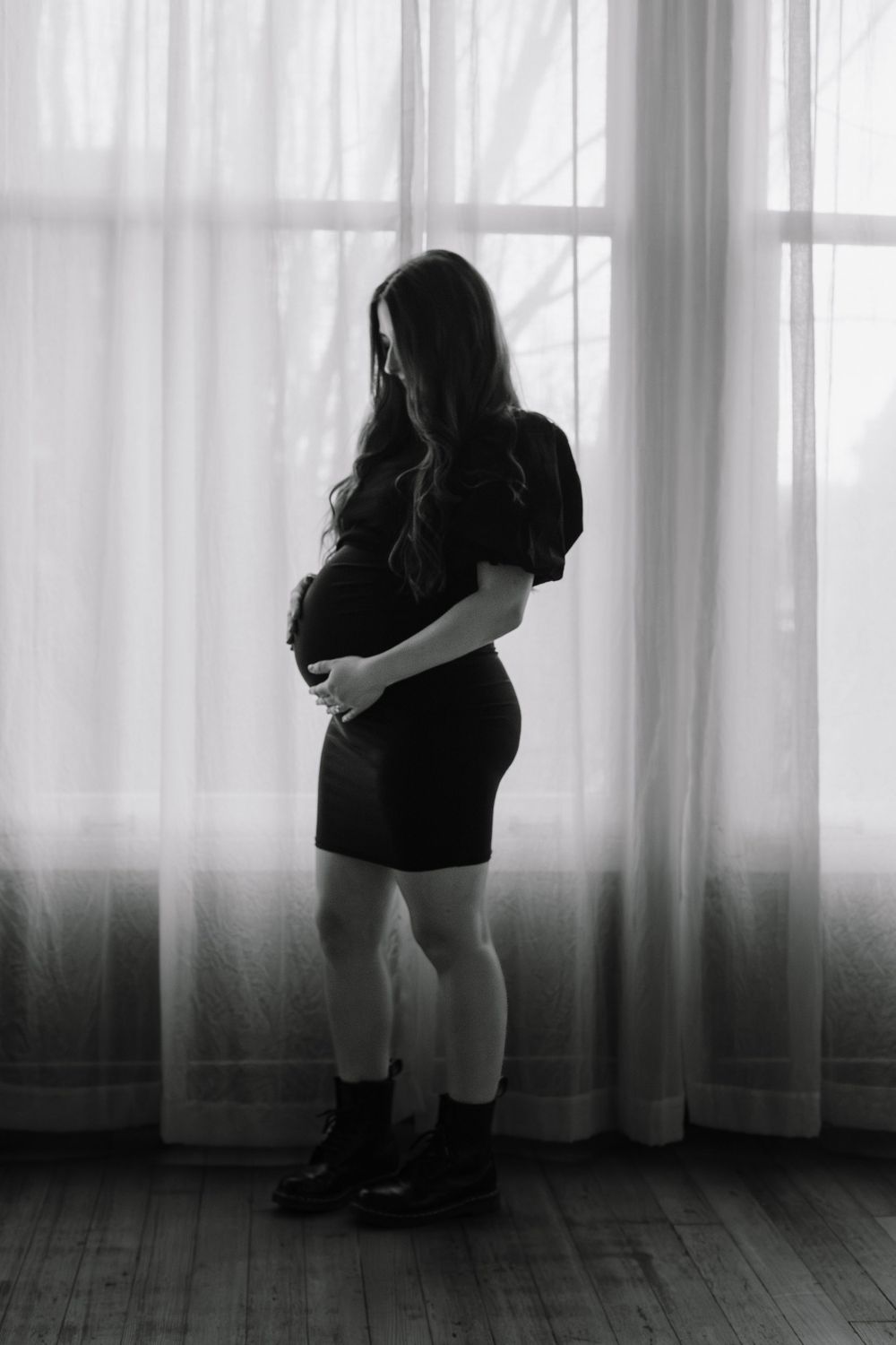 A silhouetted figure in a black dress stands contemplatively by sheer curtained windows in dramatic lighting.