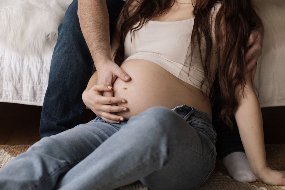 Close up of pregnant belly with hands resting gently on bare midriff while sitting in casual denim jeans.