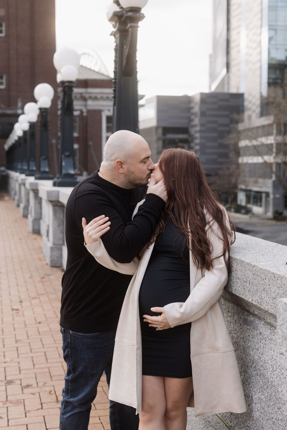 A couple shares an intimate moment on an urban walkway lined with ornate lampposts and city buildings in the background.