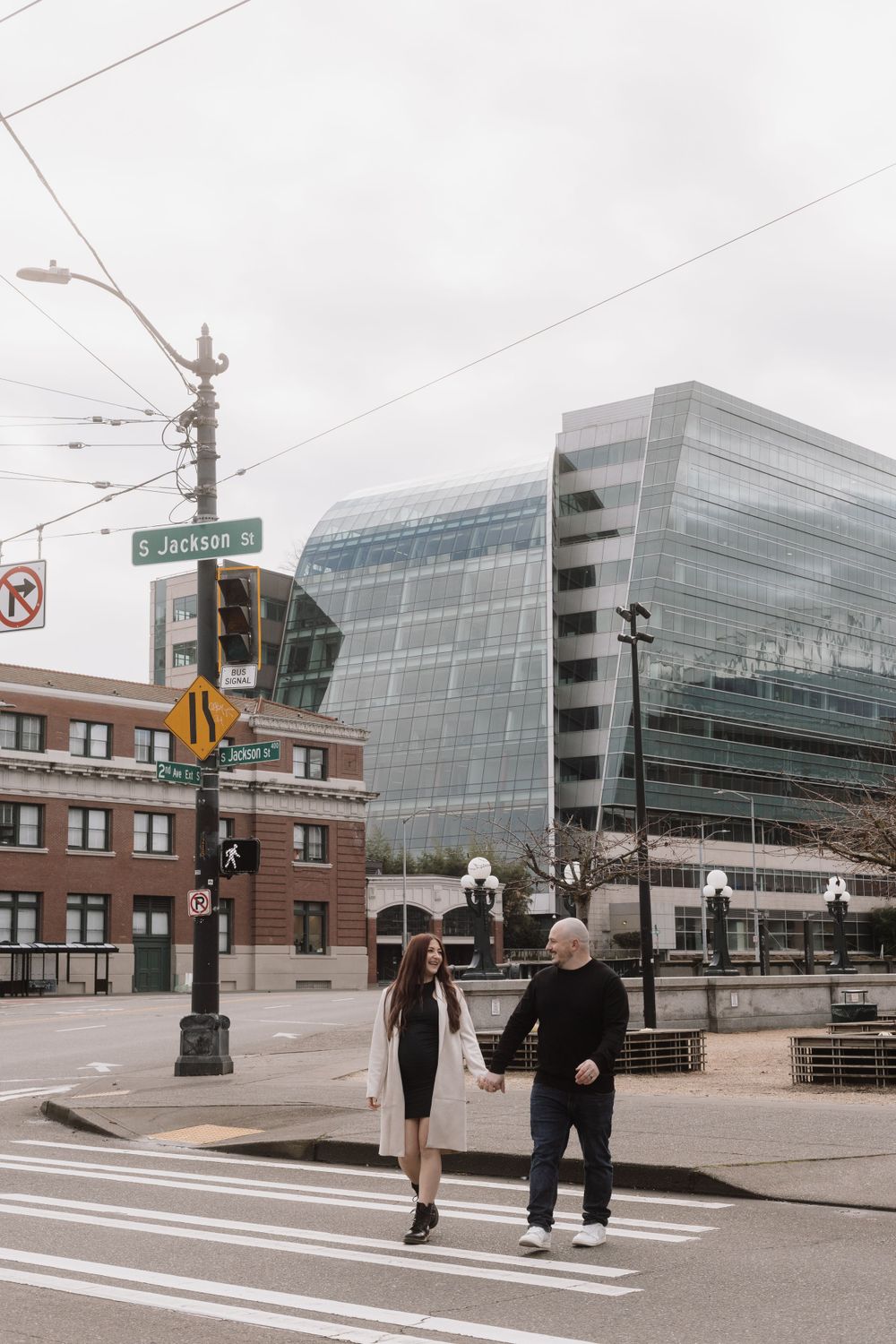 Two people crossing a street with a modern glass building and brick structure in downtown cityscape.