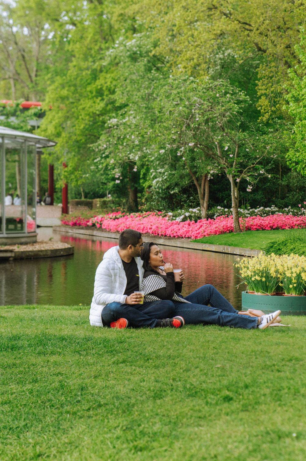 Couple relaxing on grass by pond surrounded by vibrant spring flowers and trees.