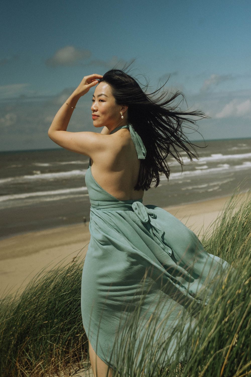 A woman in a flowing mint green dress poses on a windswept beach with dune grass.