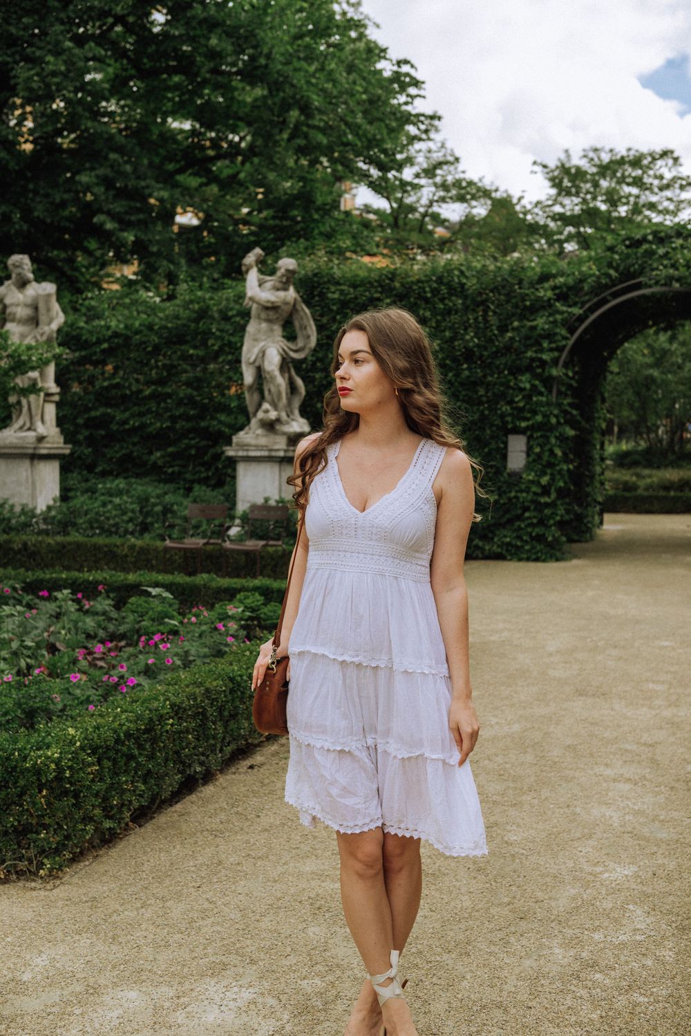 Person in white summer dress walking through a formal garden with classical statues.