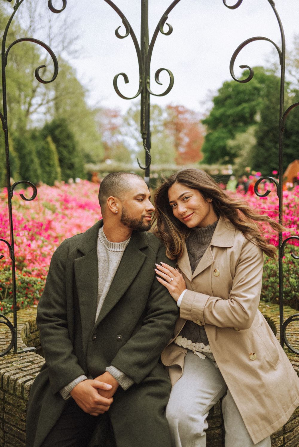 Romantic couple sharing an intimate moment by ornate garden gate with pink azaleas blooming in background.