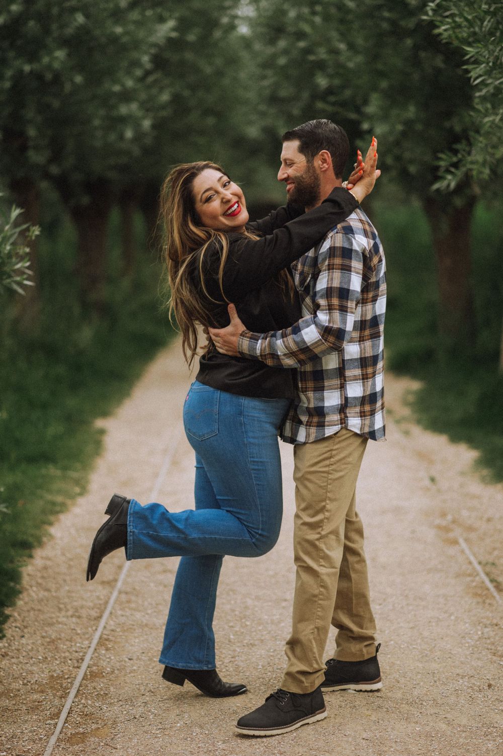 Joyful couple embracing on tree-lined dirt path during outdoor photoshoot.