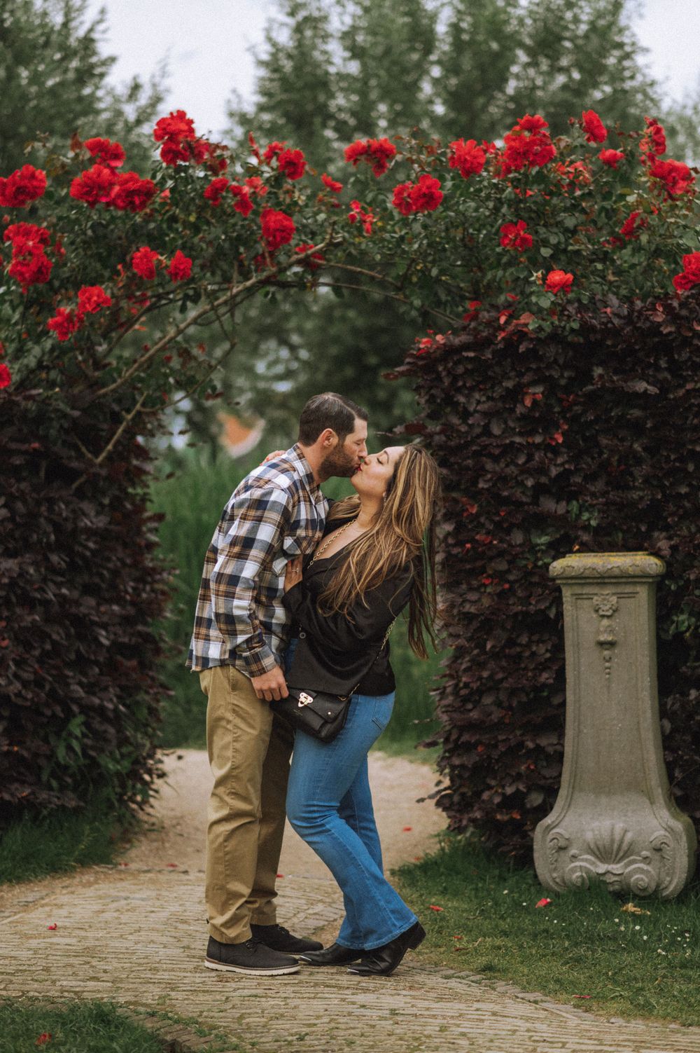 Romantic moment between couple under arch of red roses in garden path.