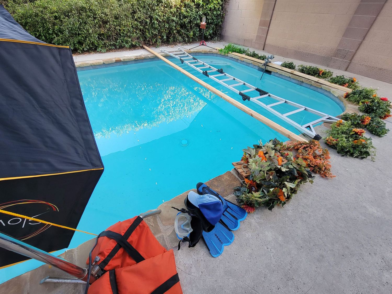 Underwater shoot- Video camera on tripod aimed at vibrant blue swimming pool with plants and pool equipment in backyard setting.