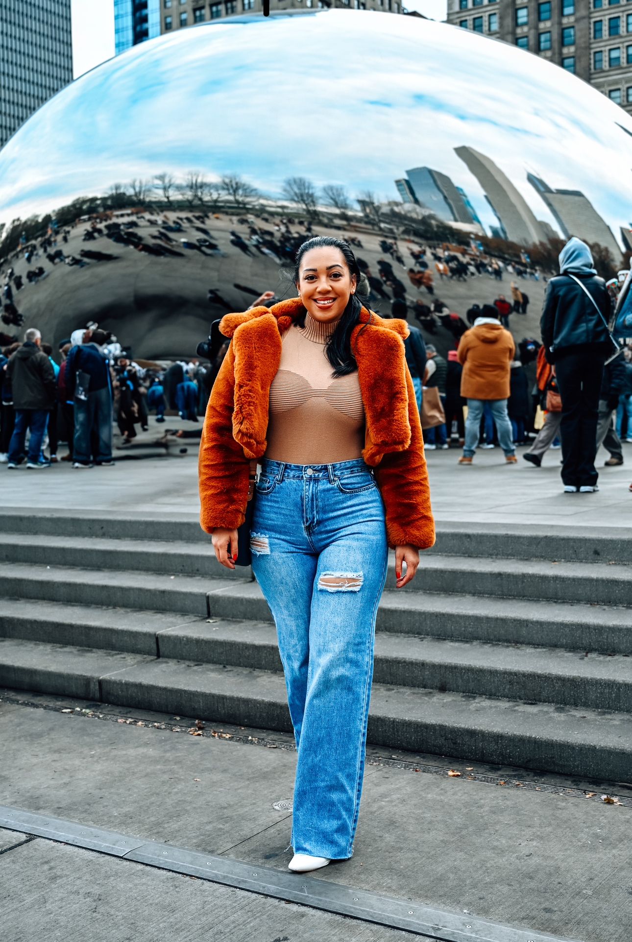 A person in an orange fur jacket and ripped jeans poses in front of the Chicago Cloud Gate sculpture on a winter day.