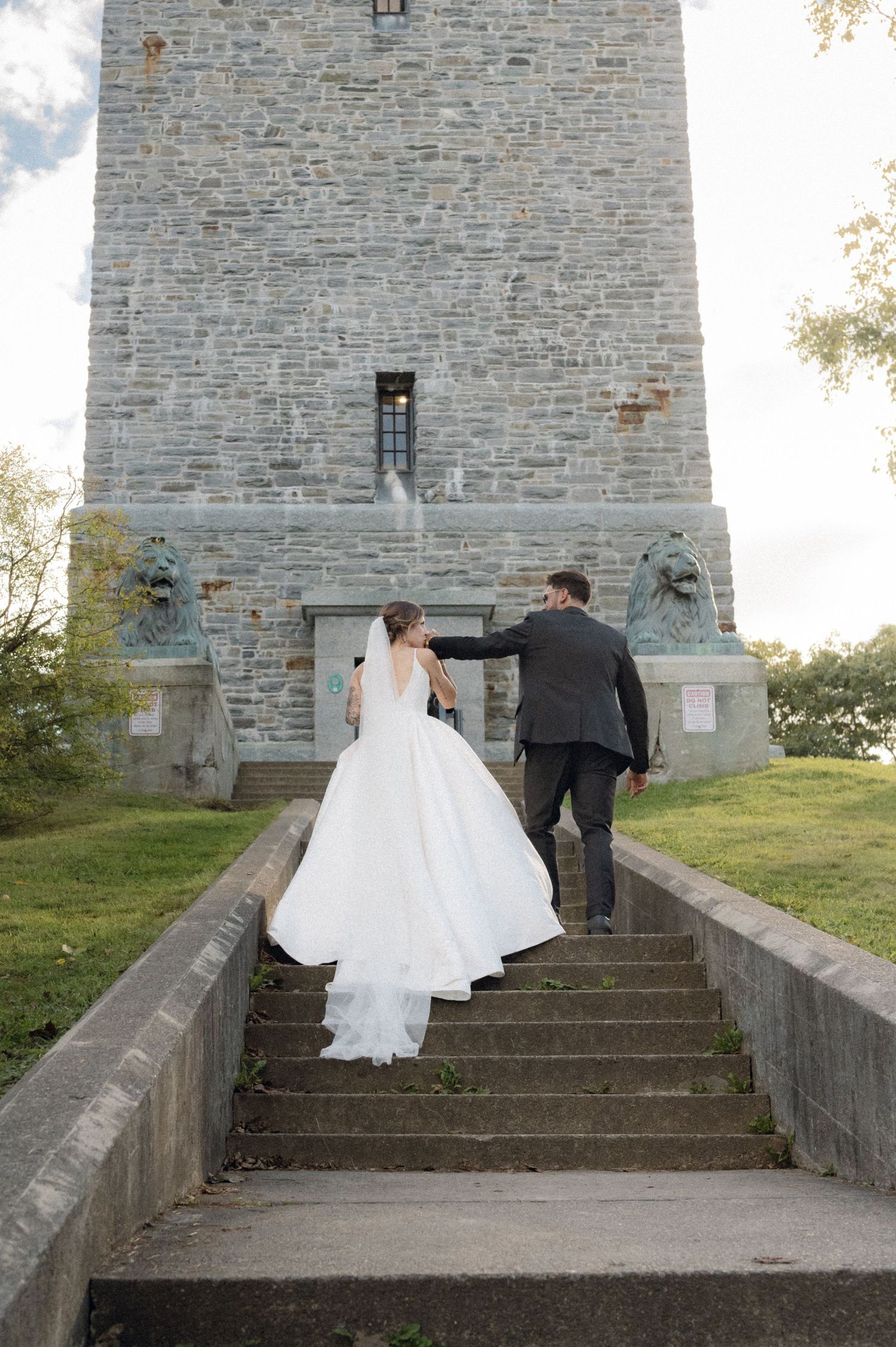 Newlyweds climbing stone steps to historic tower in wedding attire.