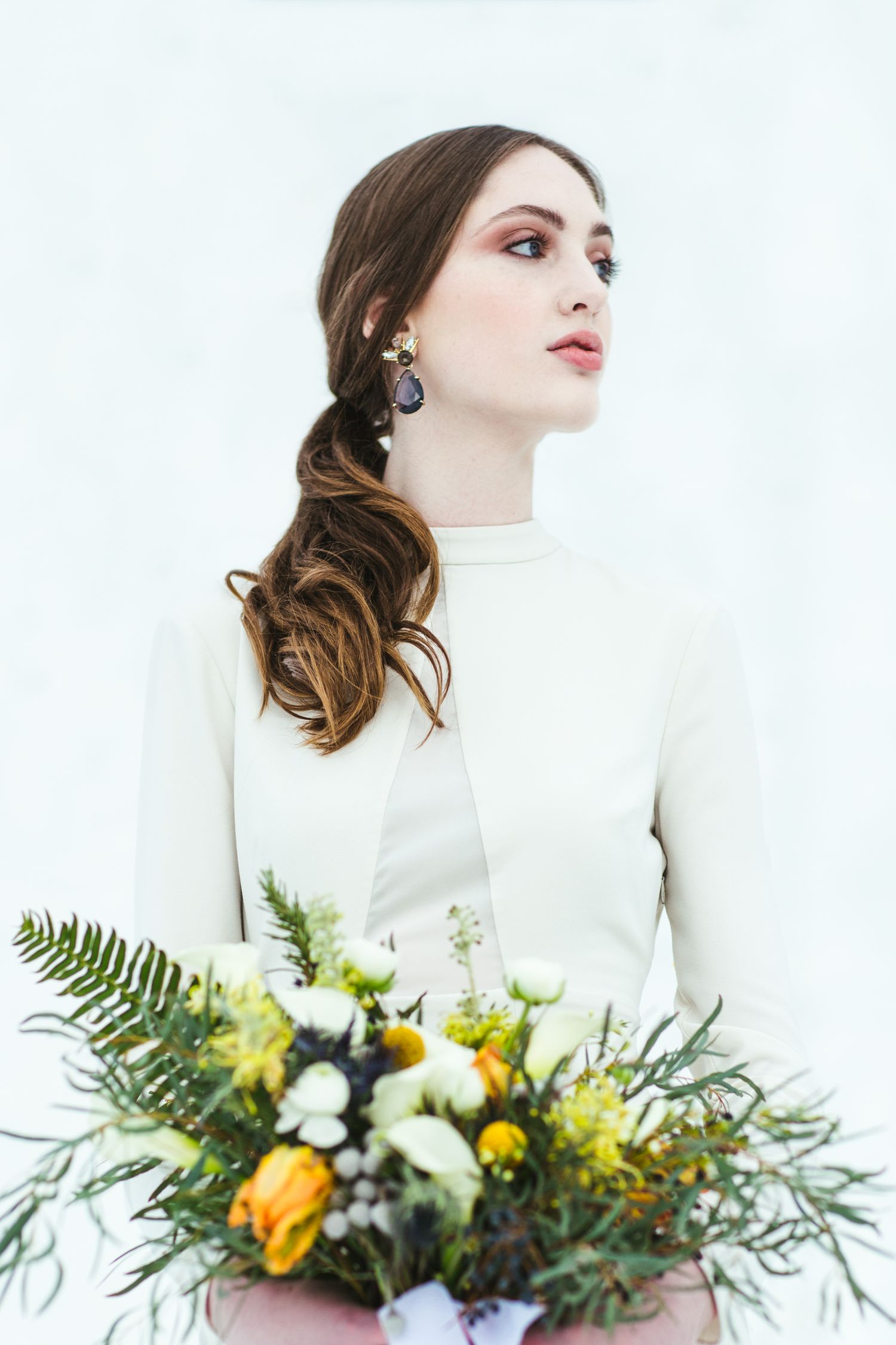 A portrait of a winter bride with elegant earrings holding a bouquet of yellow roses and ferns against a white backdrop.
