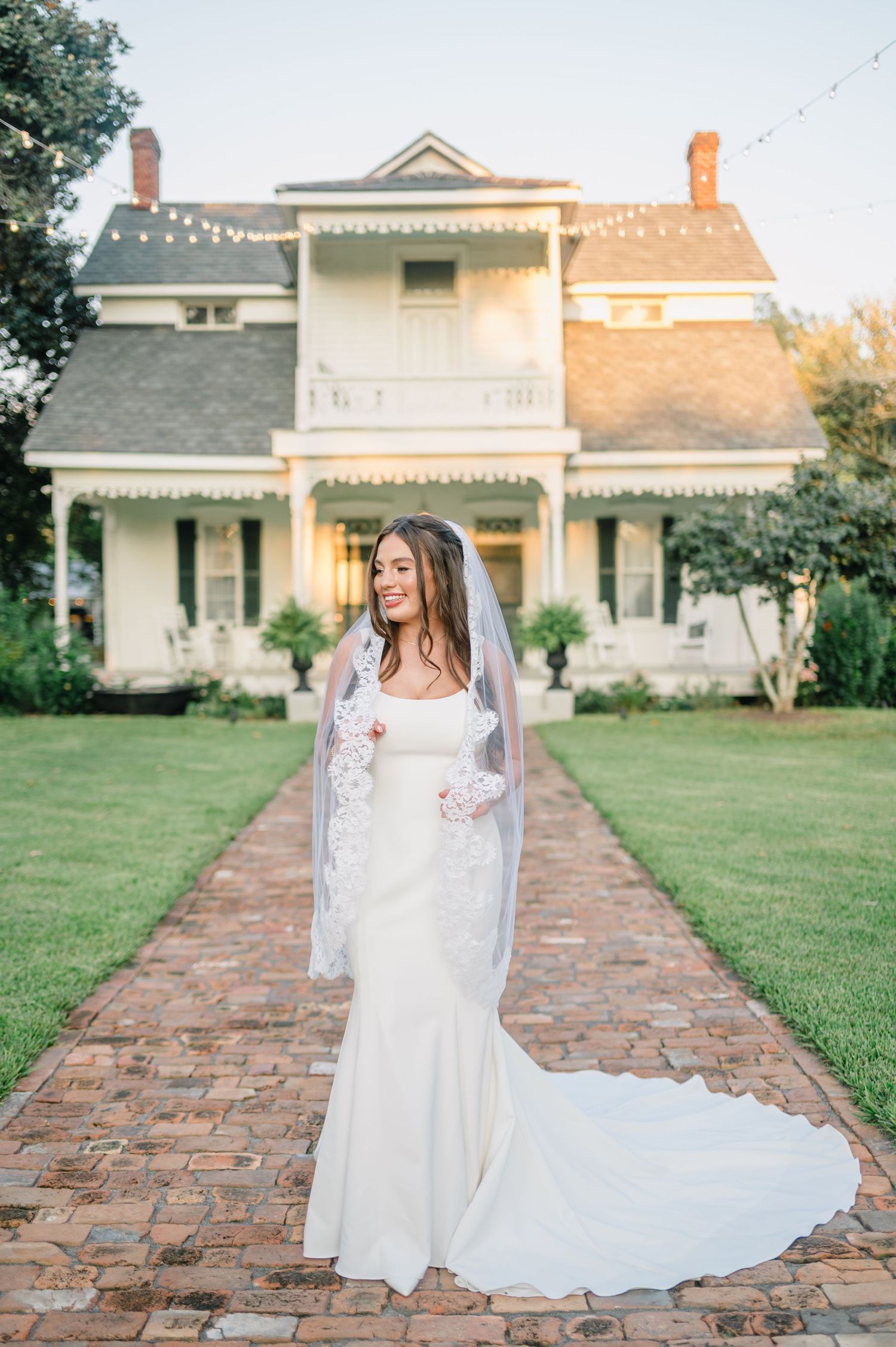 A bride in an elegant white dress with pearl-adorned veil poses outdoors in soft natural lighting.