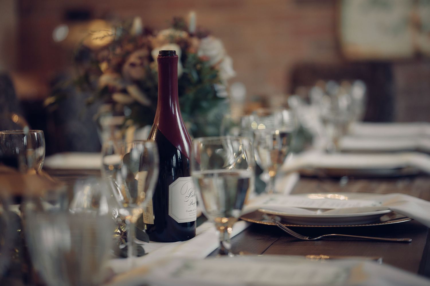 A bottle of wine sits on an elegant dinner table setting with wine glasses and place settings in soft focus.