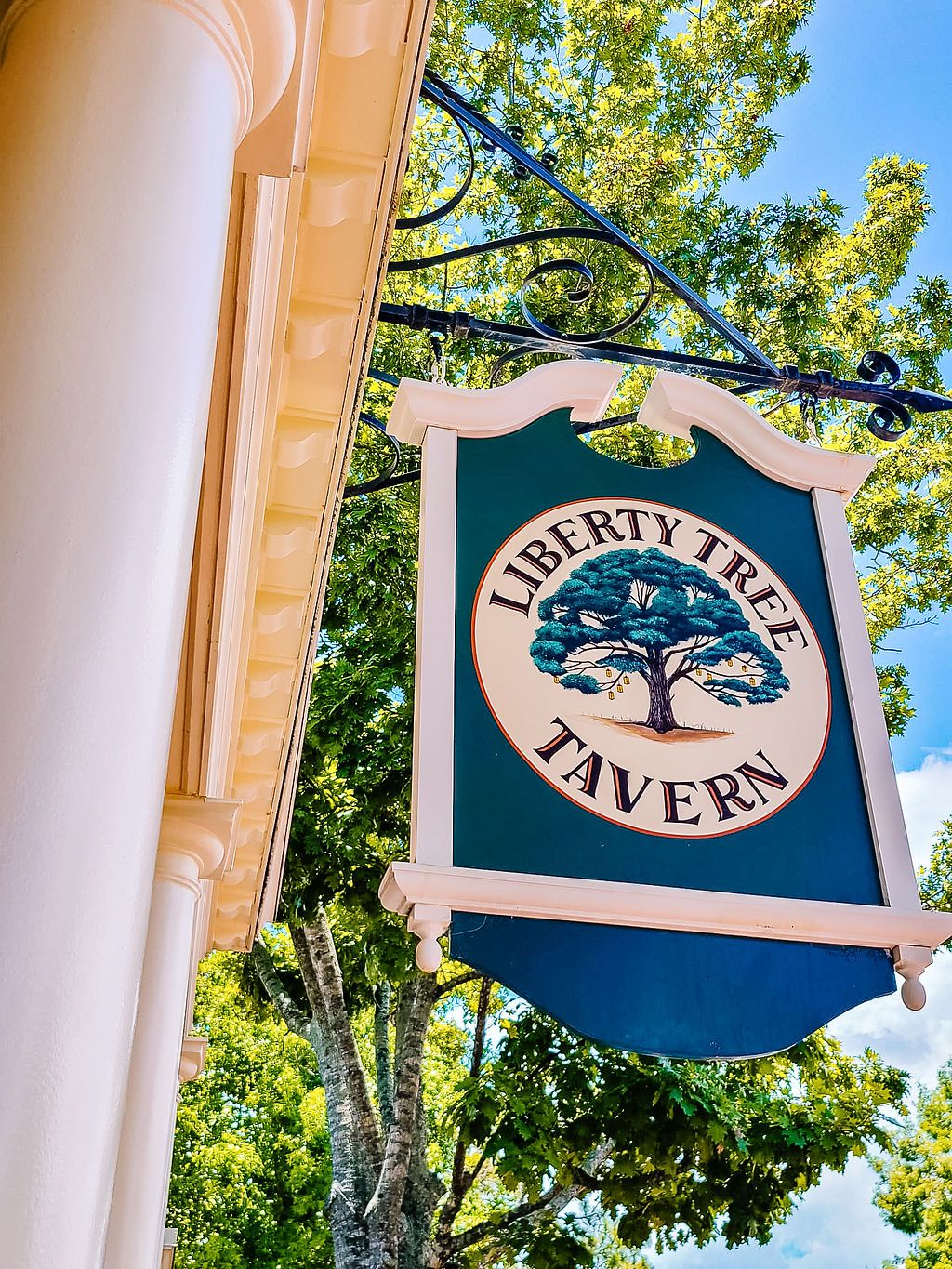 A green and white hanging tavern sign displaying a tree logo is mounted against a bright outdoor background with trees.